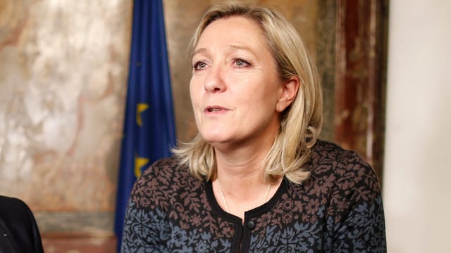 Porträt von Marine Le Pen, Blick leicht nach links, im Hintergrund eine EU-Fahne an die Wand gelehnt.