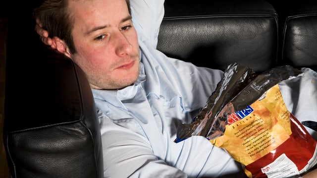 Ein Mann liegt auf einer schwarzen Couch mit einer Tüte Chips im Arm.