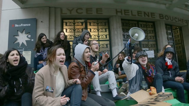 Junge Menschen sitzen auf einer Treppe und skandieren einen Sprechchor, auch ein Megafon ist zu sehen.