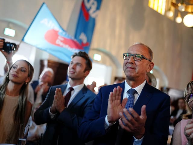 Männer klatschen in einer Halle in die Hände. AfD-Fahnen werden geschwenkt.