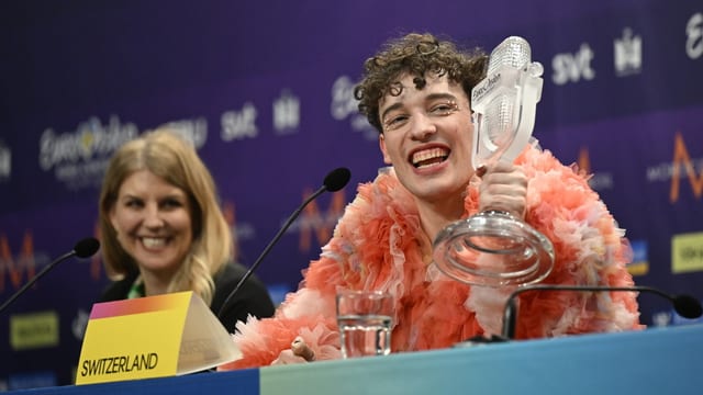 Junge Person in rosa Federrobe hält einen Pokal, sitzt neben einer Frau bei einer Pressekonferenz.