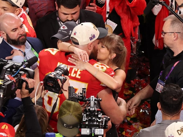 Taylor Swift und Travis Kelce küssen sich umgeben von zahlreichen Journalistinnen und Journalisten.