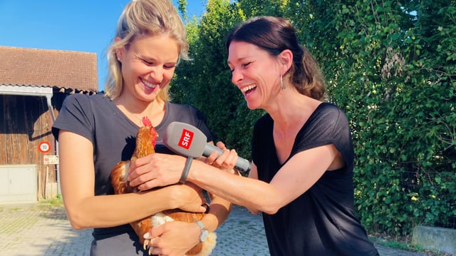 Zwei Frauen auf dem Bild. Eine hält ein Huhn in der Hand. Die andere ein Mikrofon mit dem sie das Huhn interviewt.