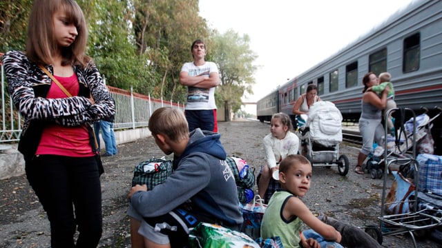 Mütter mit Kindern und einige Jugendliche stehen und sitzen neben einem Zugswaggon.