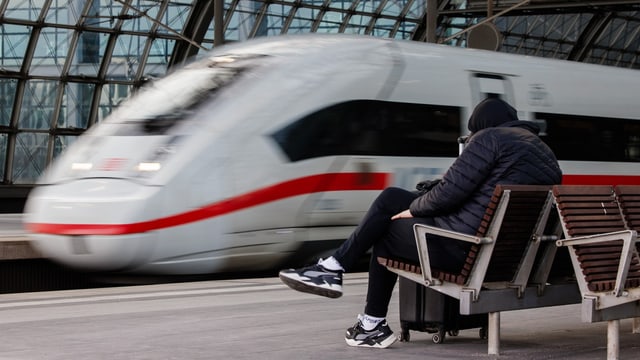 verschwommener Zug der DB, davor scharf ein Mann mit Kapuze auf einer Wartebank.