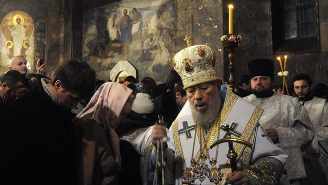 Prunkvoll gekleidete Priester tragen Kleinodien durch ein von Kerzen beleuchtetes Kloster.