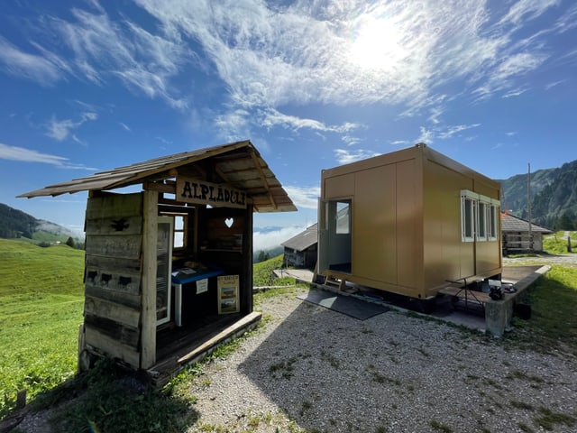 Links das Alplädeli, rechts der gelbe Container, blauer Himmel und Sonnenschein.