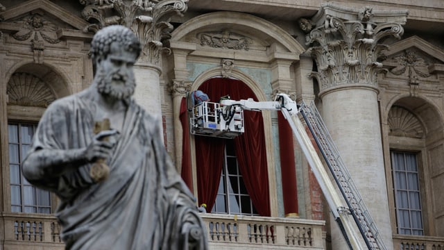 Im Vordergrund sieht man die Staue des Apostel Paulus vor dem Petersdom. Im Hintergrund montieren Menschen einen roten Vorhang vor die Balkontür.