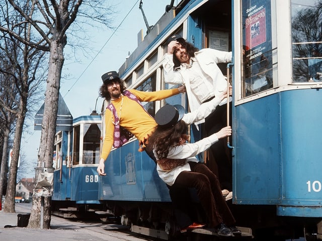 Bärtige junge Männer als Schaffner verkleidet lehen sich ein Tram hinaus. 