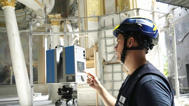 Ein junger Mann mit blauem T-shirt und Helm, darauf steht «Unesco». Er bedient ein Gerät auf einem Stativ.