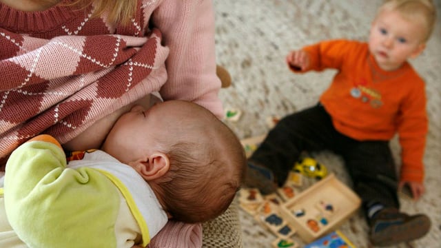 Eine Frau stillt ein Baby, dahinter sitzt ein Kleinkind auf dem Boden.