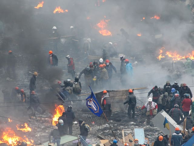 Lodernde Feuer, Menschen mit Helmen im Rauch, eine Gruppe trägt auf einer Bahre einen Verletzten davon.