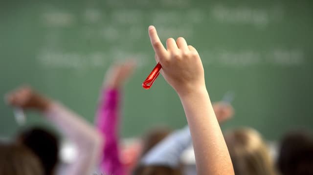 Kind streckt im Unterricht auf, man sieht nur die Hand des Kinder scharf und erahnt, dass auch andere aufstrecken.