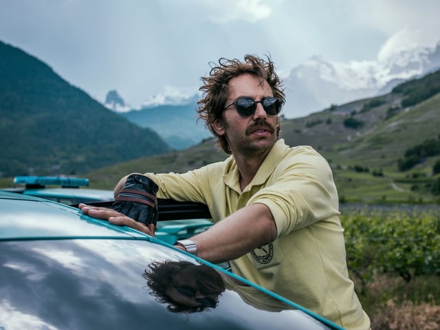 Mann mit Sonnenbrille lehnt an einem Auto, Berglandschaft im Hintergrund.