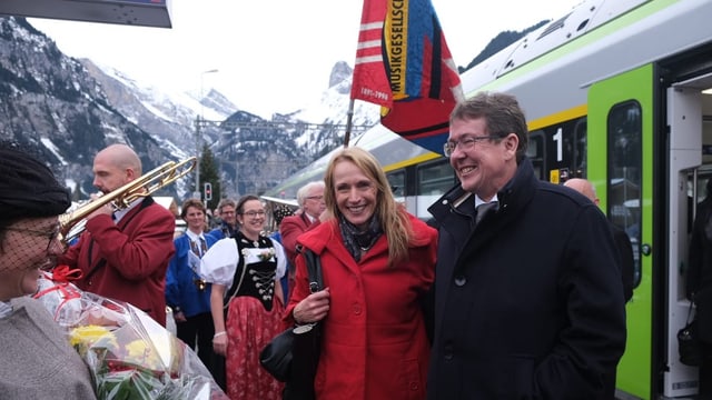 Das Ehepaar Rösti auf dem Perron, Menschen in Trachten, eine Fahne der Musikgesellschaft, im Hintergrund Berge.