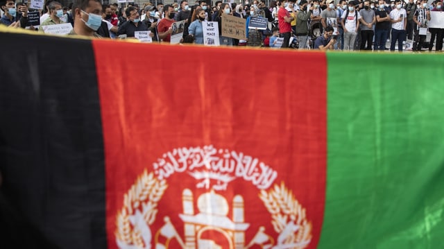 Hochgehaltene Flagge Afghanistans; im Hintergrund sind demonstrierende Personen zu sehen