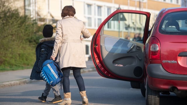 Eine Mutter steigt mit ihrem Kind aus dem Auto. Im Hintergrund sieht man ein Schulgebäude.