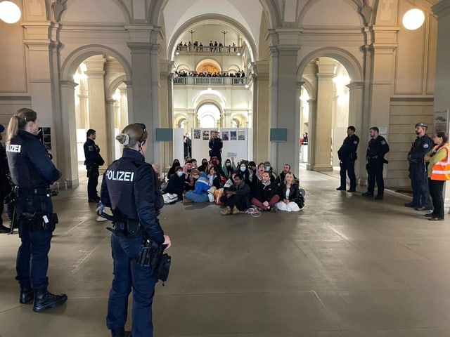 Demonstranten sitzen in einem Bahnhofsgebäude, umgeben von Polizeibeamten.