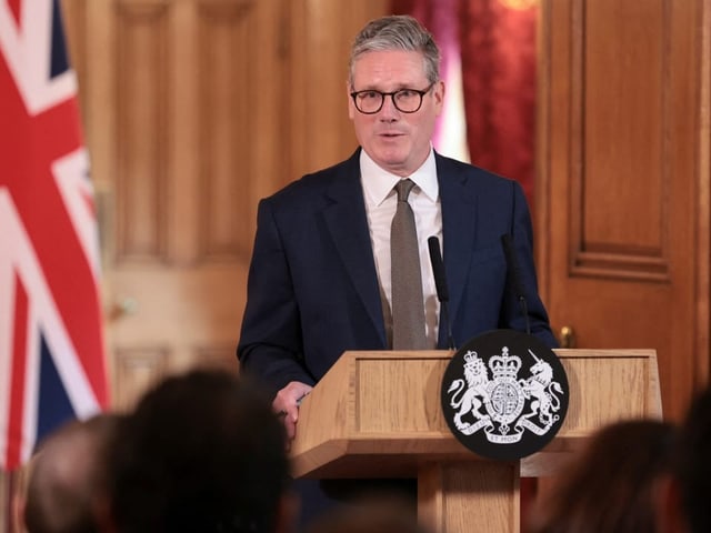 Mann im Anzug hält eine Rede hinter einem Podium vor der britischen Flagge.