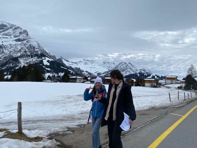 Anik hält einem Mädchen das Mikrofon hin. Sie laufen auf der Strasse, hinter ihnen ist eine Bergkette zu sehen.