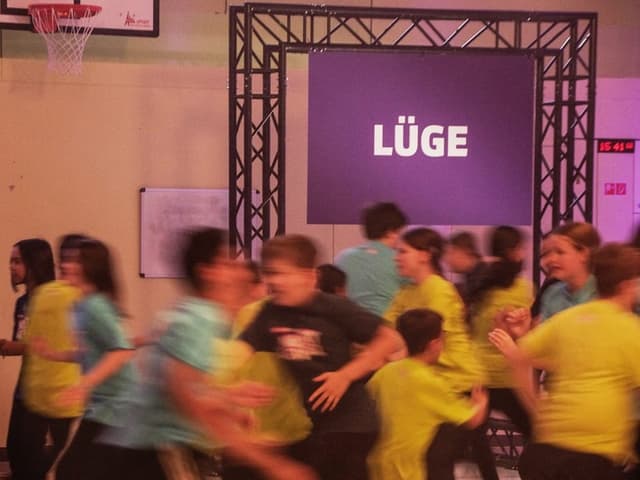 Kinder rennen durch den Raum; im Hintergrund ein Banner mit Aufschrift «Lüge»