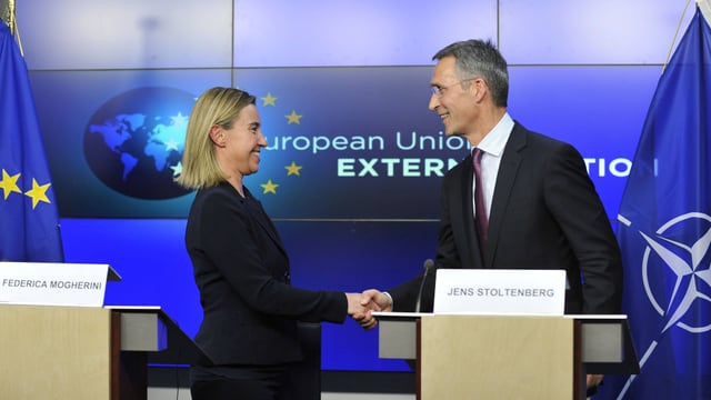 Mogherini und Stoltenberg geben sich anlässlich einer Medienkonferenz hinter ihrn Rednerpulten die Hand, im Hintergrund Nato- und EU-Flagge.