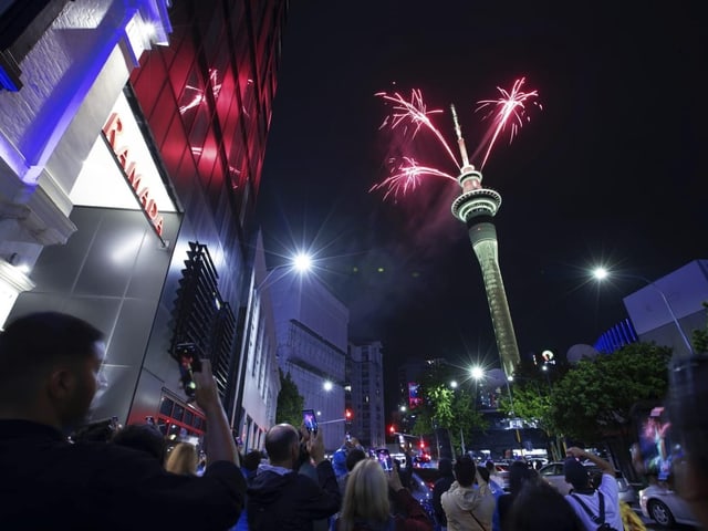 Menschen auf der Strasse schauen zum Turm hoch. Darüber leuchtet Feuerwerk.