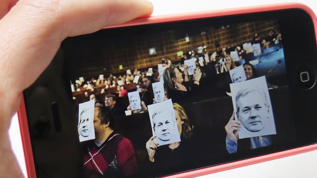 Ein Handy zeigt ein Foto, auf dem Teilnehmer einer Konferenz sich das Bild von Julian Assange vors Gesicht halten.