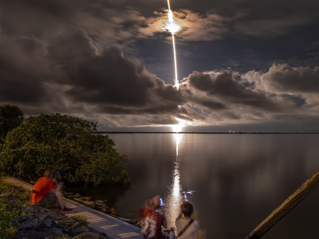 Nächtlicher Raketenstart über einer Wasserfläche mit Wolken am Himmel.