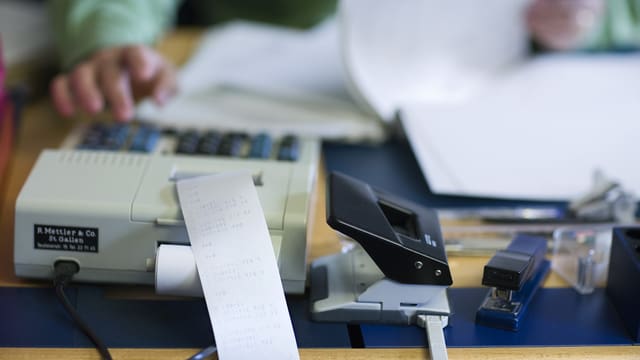 Ein Pult mit einem Papierberg und einer Rechenmaschine, auf der eine Hand etwas eintippt.