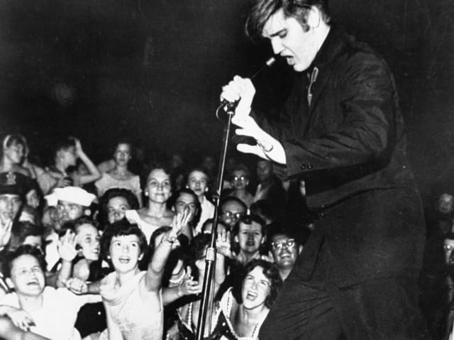 Elvis Presley tanzt auf der Bühne, davor stehen kreischende Fans