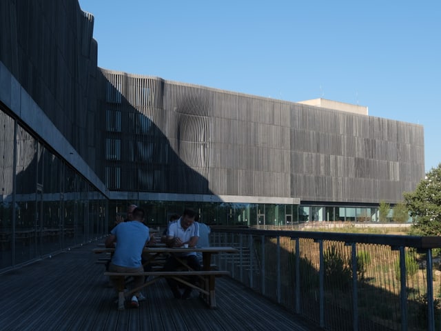 Menschen sitzen an Picknicktischen auf einer Terrasse vor einem modernen Gebäude.