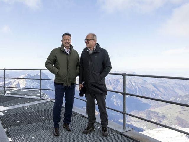 Zwei Männer in Winterkleidung stehen vor einem Bergpanorama.