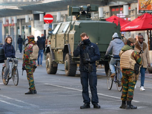 Am Strassenrand abgestelltes gepanzertes Militärfahrzeug mit zwei Soladten davor, in der Strasse wenige Passanten, zwei davon auf Velos