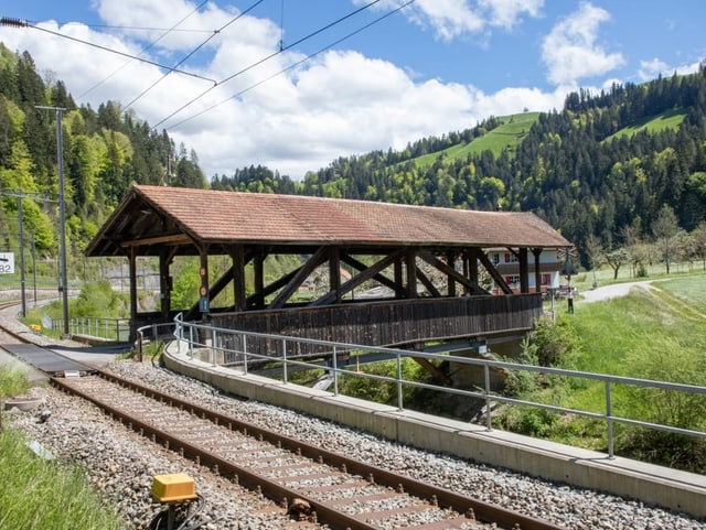 Gedeckte Holzbrücke am Bahngleis über einen kleinen Bach.