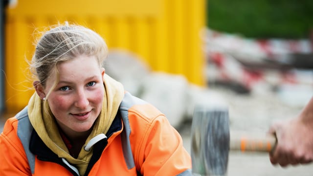 Die junge Strassenbauerin Katja schaut in die Kamera. Hinter ihr die Baustelle. 