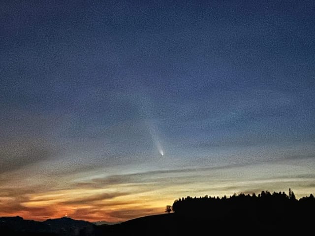 Nachthimmel mit Komet über bewaldeten Hügeln im Sonnenuntergang.