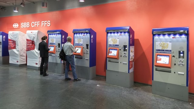 Mehrere Billettautomaten der SBB stehen in einer Reihe. Davor stehen zwei Reisende.