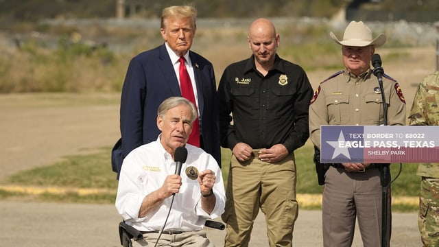 Greg Abbott spricht bei einem Anlass in ein Mikrofon. Er sitzt in seinem Rollstuhl vor Ex-US-Präsident Donald Trump.