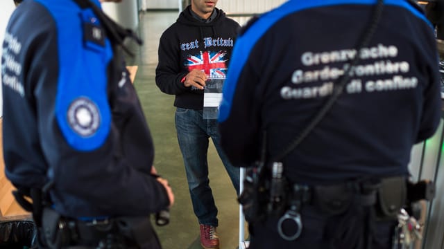 Zwei Grenzwächter in Uniform von hinten fotografiert stehen vor einem Mann, der einen Pullover mit der Flagge des Vereinigten Königreichs trägt.