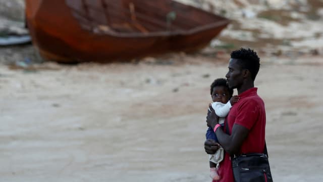 Junger Mann hält ein Kind auf dem Arm, im Hintergrund ein gestrandetes Boot