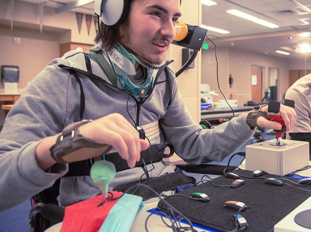 Ein körperlicher behinderter Junger Mann, dessen Hände an Joysticks und Knöpfen fixiert sind, vor ihm weitere Knöpfe.