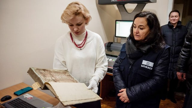 Eine Frau hält ein altes Buch in der Hand, eine weitere Frau steht daneben. Im Hintergrund steht ein Scanner.