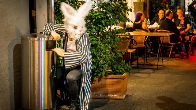 Links ein metallener Mülleimer, ein Mensch mit Hasenkopf-Maske kniet daneben. Im Hintergrund sitzen Menschen draussen