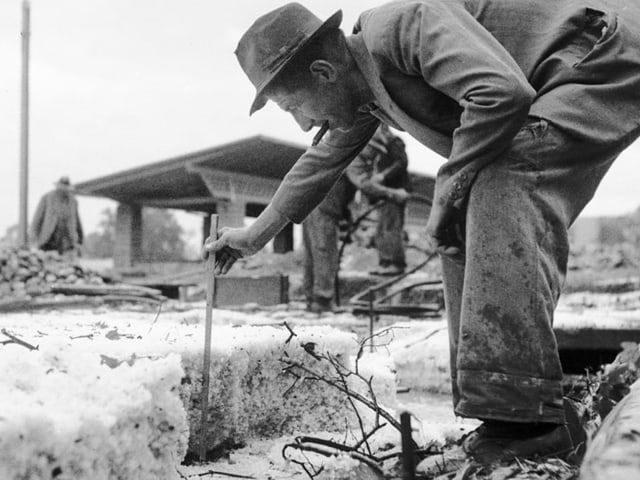 Mann misst Schnee mit einem Stock während anderer Männer im Hintergrund arbeiten.