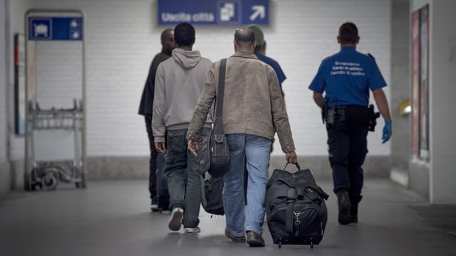 Eine Gruppe Migranten von hinten geht zusammen mit einem Grenzwächter eine Bahnhofsuntrführung entlang.