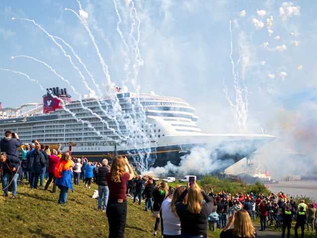 Ein Kreuzfahrtschiff, Zuschauende bejubeln die Jungfernfahrt, Feuerwerke werden gezündet. 