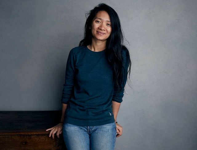 Zhao is leaning against a gray wall and smiling at the camera.