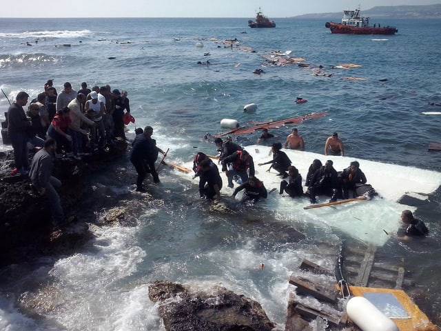 Flüchtlinge halten sich an einem Wrackteil fest und treiben im Wasser. Menschen an Land versuchen zu helfen. 