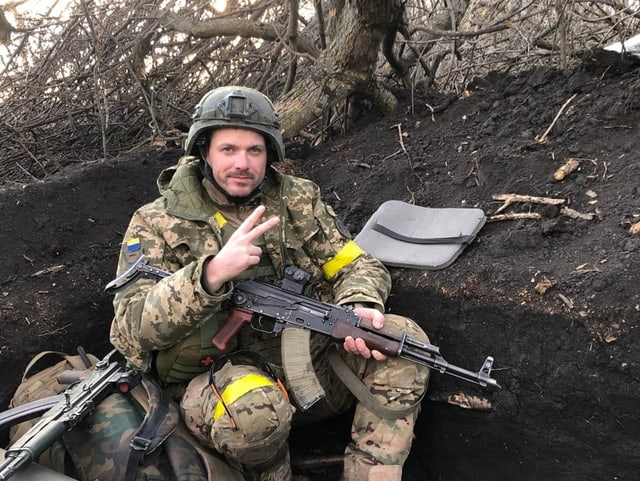 Jérôme sitzt mit Gewehr im Schützengraben und zeigt das Victory-Zeichen 